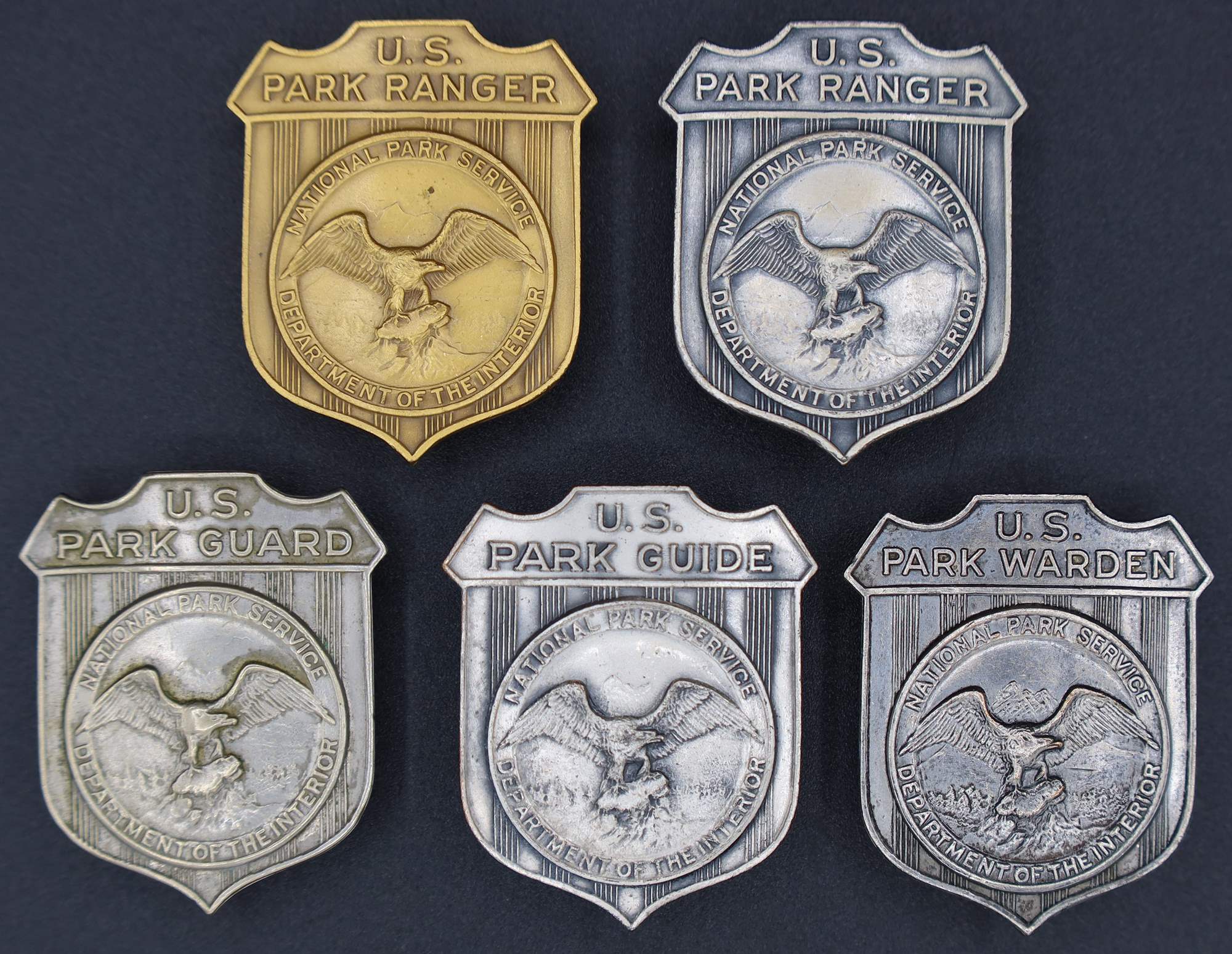 Shield-shaped badges with left facing eagle in the middle. Gold and silver marked U.S. Park Ranger, others marked U.S. Park Guide, U.S. Park Guard, and U.S. Park Warden.